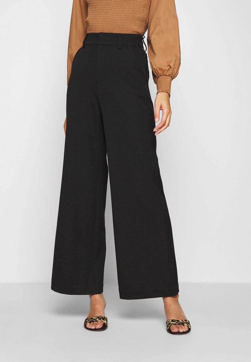 Pantaloni neri a gamba larga realizzati in tessuto morbido, dotati di vita alta e vestibilità sartoriale. Abbinati a sandali piatti con stampa animale.