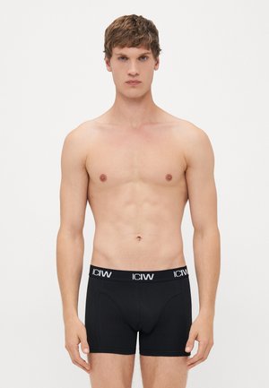 Junger Mann steht shirtlos in schwarzen Boxershorts mit weißem "ICIW"-Logo am Bund vor einem schlichten hellen Hintergrund.