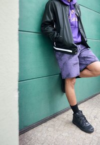 Černá bomber bunda s bílými pruhy, fialová mikina a fialové šortky s tie-dye. Černé vyšší tenisky a tmavé ponožky dotvářejí tento outfit.