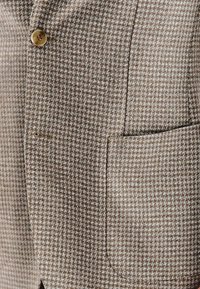 Brązowa marynarka w wzór houndstooth, z uporządkowanym krojem, pojedynczym zapięciem na guzik i kieszenią na piersi. Miękka faktura z klasycznym wzorem.