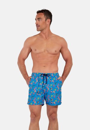 Man wearing blue swim shorts with colorful kite patterns, hands in pockets, looking to the side against a plain background.