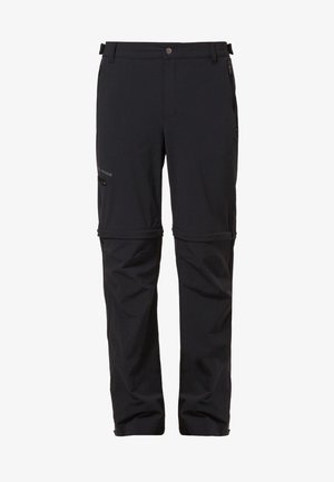 Man met een donkergrijze longsleeve, zwarte cargobroek en bruine wandelschoenen, staande tegen een effen lichte achtergrond.