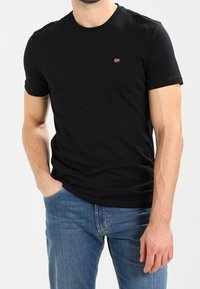 Maglietta nera in cotone con colletto rotondo e maniche corte. Presenta un piccolo logo rosso e bianco sul petto. Indossata con jeans in denim blu.