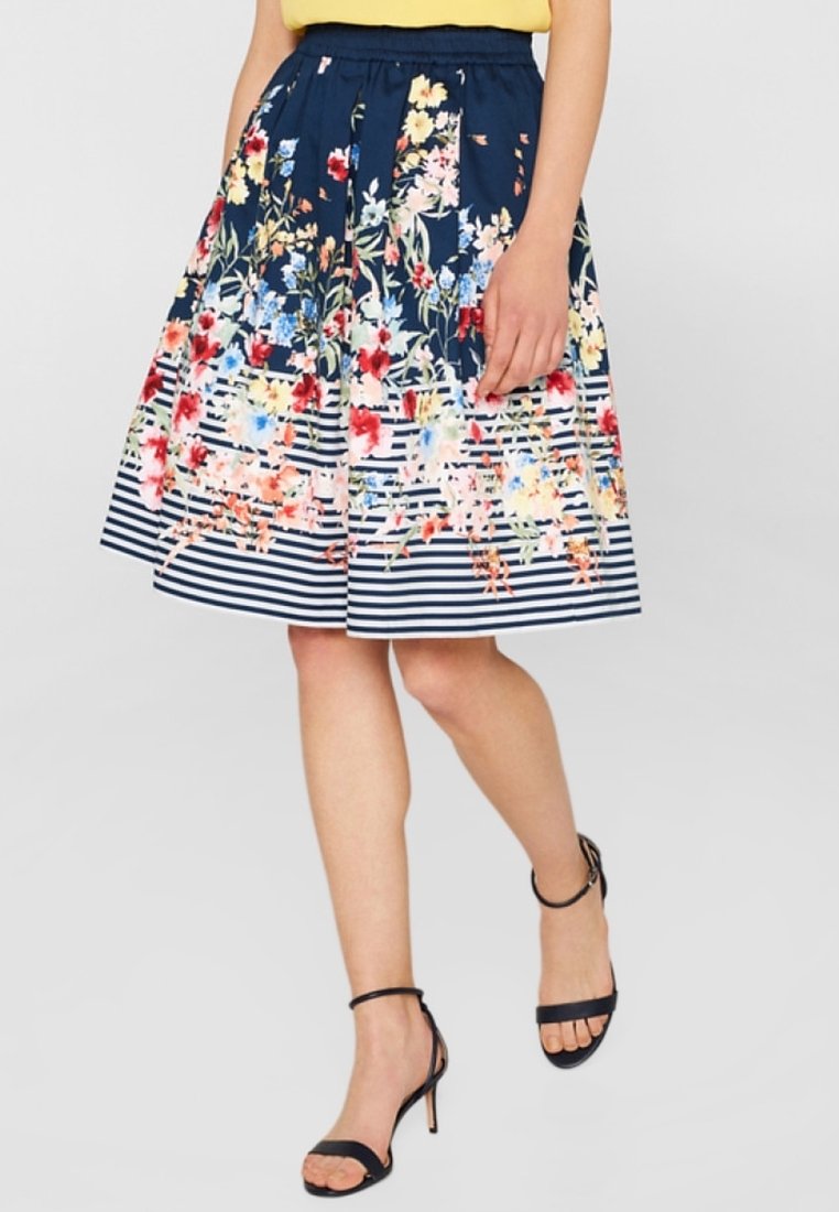 Femme portant une jupe bleu marine jusqu'aux genoux avec un motif floral et des rayures blanches horizontales, associée à des sandales noires à talons.