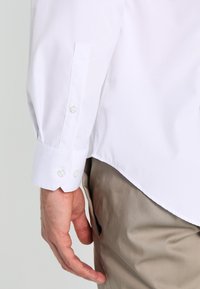 Hand van een man naast de zak van beige broek, gekleed in een witte longsleeve shirt met een dichtgeknoopte manchet, tegen een effen lichte achtergrond.