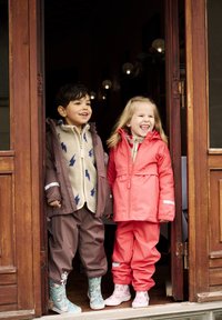 Två barn står i en dörröppning. Pojken har på sig en brun jacka med fleecefoder och blå mönster, tillsammans med byxor och mönstrade stövlar. Flickan har på sig en ljusrosa jacka och byxor, med ljusrosa blommiga stövlar.