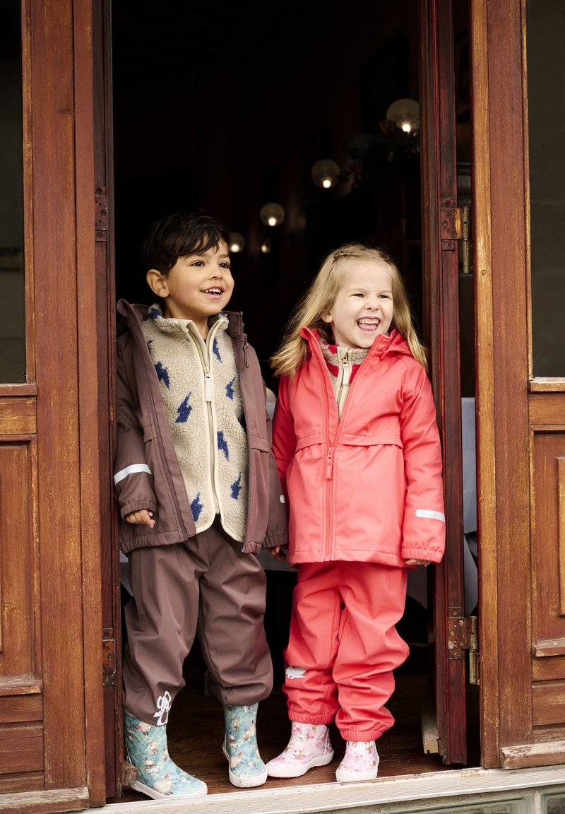 Två barn står i en dörröppning. Pojken har på sig en brun jacka med fleecefoder och blå mönster, tillsammans med byxor och mönstrade stövlar. Flickan har på sig en ljusrosa jacka och byxor, med ljusrosa blommiga stövlar.