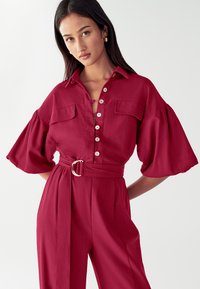 Raudona lino marškinių suknelė su mygtukų užtrauktuku, dviem krūtinės kišenėmis, plačiomis rankovėmis, diržu juosmeniui su sidabriniu užsegimu, lygia tekstūra.
