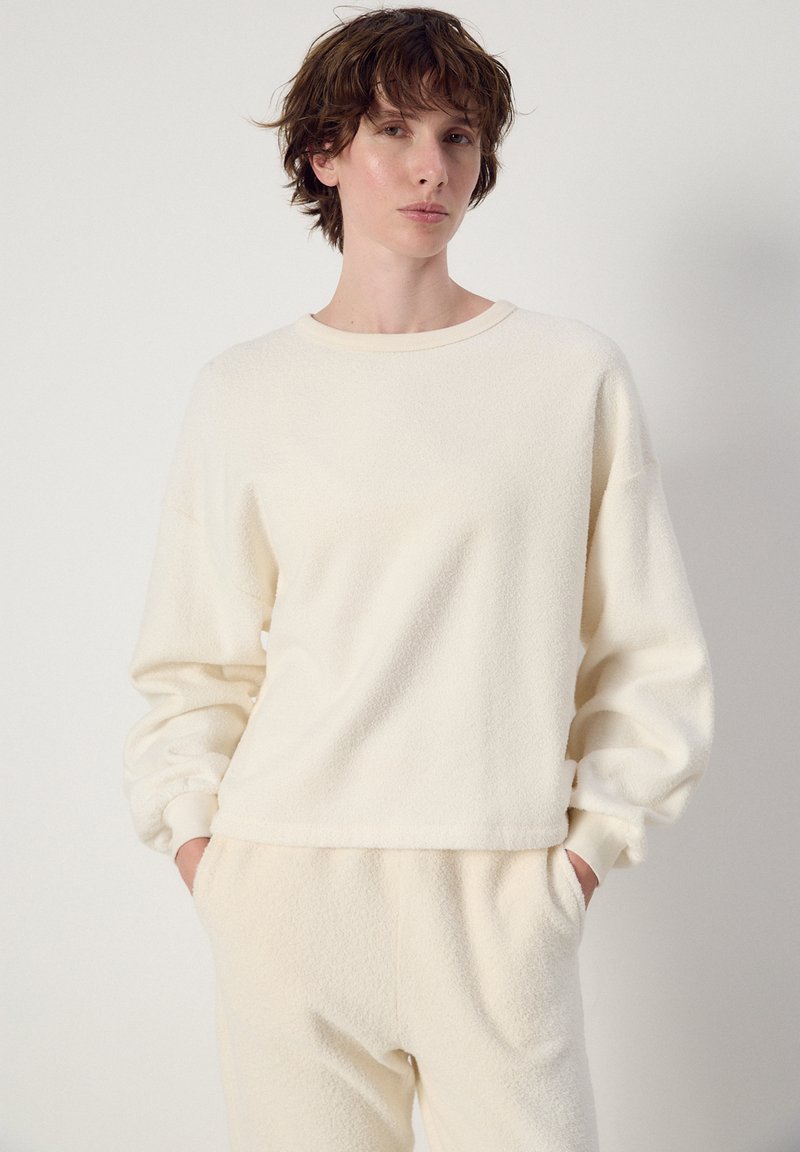 Person med kort krøllet hår iført en blød, langærmet cremefarvet sweater og matchende bukser med hænderne i lommerne mod en ensartet baggrund.