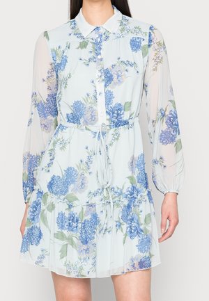 Robe fleurie bleu clair avec manches longues transparentes, devant boutonné, taille cintrée par un cordon et ourlet volanté portée par une personne debout.