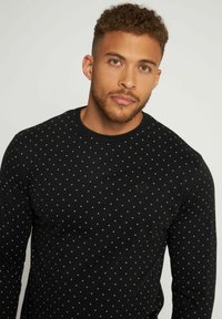 Chemise noire à manches longues ornée de petits pois blancs, avec un col rond et un tissu texturé comportant un logo discret sur la poitrine.