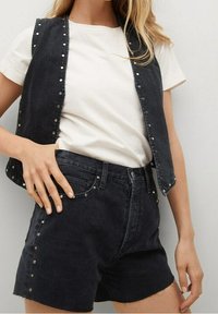 Chaleco de mezclilla negro con tachuelas plateadas y una camiseta blanca; combinado con shorts de mezclilla negra, que tienen detalles de tachuelas a lo largo de los lados.