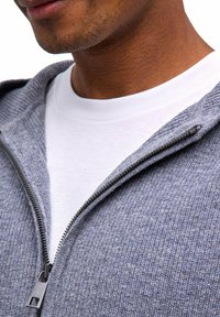 Grauer Kapuzenpullover mit Reißverschluss aus strukturiertem Stoff, ausgestattet mit einem silbernen Reißverschluss und einem weißen T-Shirt mit Rundhalsausschnitt darunter. Nahaufnahme des Halsausschnitts.
