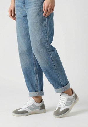 Person trägt umgeschlagene blaue Jeans und weiße Turnschuhe mit grauen Wildledereinsätzen, steht vor einem einfarbigen hellen Hintergrund.