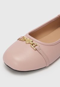 Sapatos planos rosa pálido com bico arredondado, detalhe em metal dourado com forma de coração, textura de couro suave e sola preta em contraste.