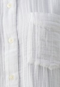 Chemise blanche à boutons en tissu léger avec une texture légèrement froissée, dotée d'une poche poitrine et de boutons ronds.