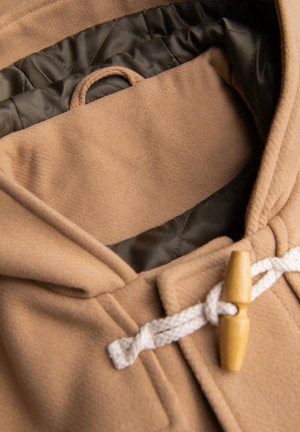 Beige Kapuzenjacke mit strukturiertem Außenmaterial, ausgestattet mit einem dunkelgrünen Innenfutter, einem Holzverschluss und einer Schlaufe zum Aufhängen.