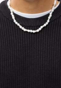Collar de perlas con cuentas blancas de acabado brillante, intercaladas con pequeños acentos metálicos, llevado sobre un suéter texturizado oscuro.