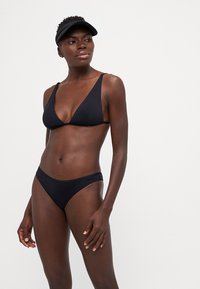 Juodas bikini su trikampiu viršumi ir žemu apatine dalimi, pagamintas iš lygaus audinio. Dėvimas su juodu vizoriu, turinčiu minimalistinį dizainą.