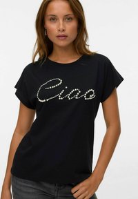 Jeune femme portant un t-shirt noir avec le texte "Ciao" orné de perles, associé à un jean gris foncé, se tenant contre un fond uni.