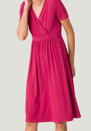 Mujer con vestido cruzado de manga corta, largo hasta la rodilla, de color rosa intenso, con cintura ajustada y pliegues suaves sobre fondo gris claro.