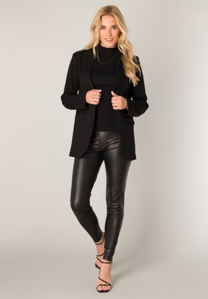 Blonde woman wearing a black blazer, black turtleneck, high-waisted black leather pants, and black strappy heels, standing confidently.