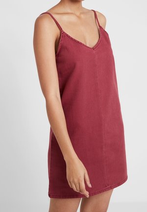 Kvinde iført ærmeløs, V-hals, dybrød afslappet kjole med tynde stropper, stående mod en ensfarvet lys baggrund.