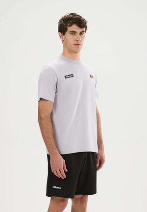 Ein junger Mann steht und trägt ein hellviolettes ellesse T-Shirt und schwarze ellesse Shorts vor einem einfarbigen hellen Hintergrund.