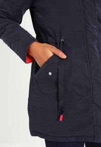 Námořnická puffer bunda s nastavitelným pasem, elastickými manžetami, postranními kapsami a kontrastní červenou podšívkou. Hladká textilie a minimalistický design.