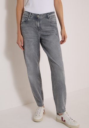 Personne portant un jean gris large et des baskets blanches avec un logo rouge, se tenant contre un mur clair uni.