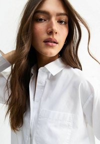 Chemise blanche à boutons en coton avec un col, une poche poitrine unique et une coupe légèrement décontractée. Texture lisse et apparence nette.