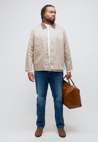 Άνδρας με πλεξούδες, φοράει μπεζ μπουφάν, λευκό πουκάμισο, μπλε τζιν, καφέ μποτάκια, κρατώντας καφέ δερμάτινη διπλή τσάντα, στέκεται μπροστά σε λευκό φόντο.