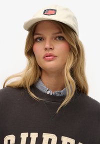 Young woman with long blonde hair wears a beige cap with an American flag patch, a black sweatshirt, and a blue checkered shirt underneath.