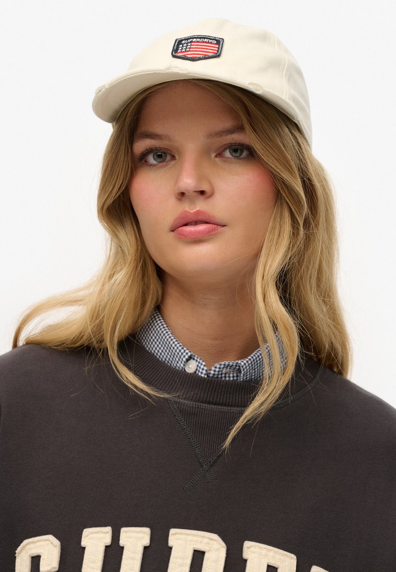 Ung kvinde med langt blondt hår bærer en beige kasket med et amerikansk flag-patch, en sort sweatshirts og en blåternet skjorte under.