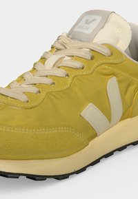Baskets de sport jaune et crème avec lacets à l’avant, logo blanc "V" sur le côté et semelle crème avec bande de roulement noire.