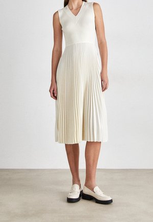 Robe midi blanche sans manches avec col en V et jupe plissée, portée avec des mocassins blancs épais sur un sol neutre et un fond uni.