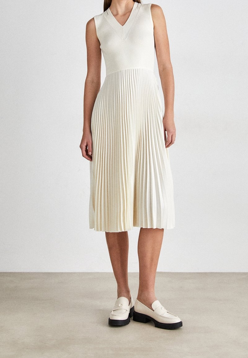 Robe midi blanche sans manches avec col en V et jupe plissée, portée avec des mocassins blancs épais sur un sol neutre et un fond uni.