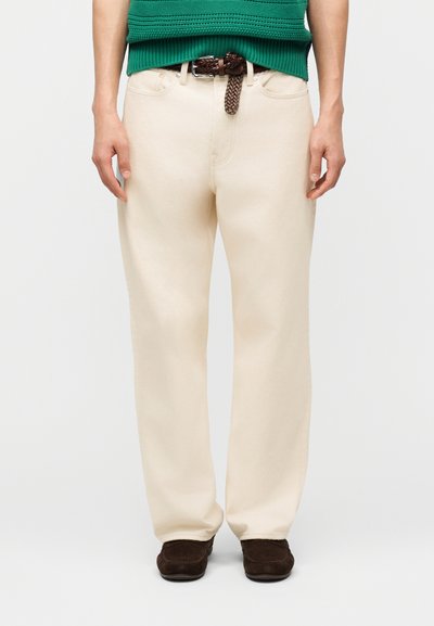Homme portant un pantalon beige à jambes droites, une ceinture tressée marron, un haut en maille vert et des mocassins marron foncé, debout devant un fond uni.