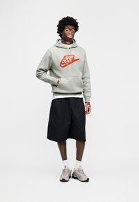 Γκρι φούτερ με κουκούλα με λογότυπο "JUST DO IT" σε κόκκινο, συνδυασμένο με μαύρες βερμούδες. Το μοντέλο φοράει ανοιχτογκρι αθλητικά παπούτσια με λευκές κάλτσες. Μινιμαλιστικό φόντο.