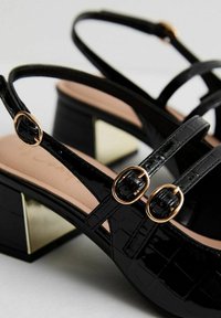 Svarta lackläderssandaler med två remmar och guldtonade spännen samt en geometrisk blockklack med metalliska detaljer.