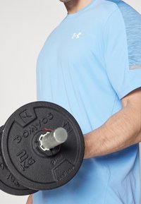 Ljusblå träningsskjorta med korta ärmar, med en texturerad axelaccent. Svart viktplatta med ett silverhandtag som hålls i ena handen.