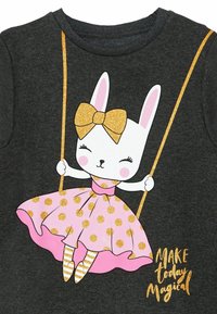Graues Baumwoll-T-Shirt mit einer Illustration eines Kaninchens auf einer Schaukel, gekleidet in einem pinken Kleid mit goldenen Punkten und einer glitzernden Schleife.