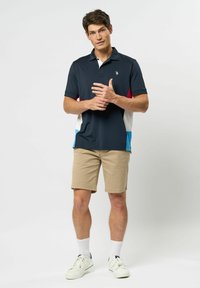 Mornarska polo majica s crvenim, bijelim i plavim bočnim panelima, uparenim s bež šortsevima. Model nosi bijele tenisice i čarape, stojeći s rukama sklopljenim.