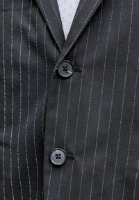 Μαύρο σακάκι με ρίγες, κουμπωμένο πάνω σε λευκό υφασμάτινο πουκάμισο, με δύο μεγάλα μαύρα κουμπιά και λεπτομέρειες στον γιακά.