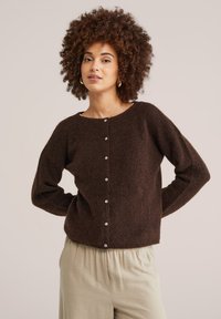 Sieviete ar cirtainiem matiem, valkā zelta stīpveida auskarus, tumši brūnu piepogātu kardiganu un gaiši beigzilas bikses, pozējot ar rokām aiz muguras.