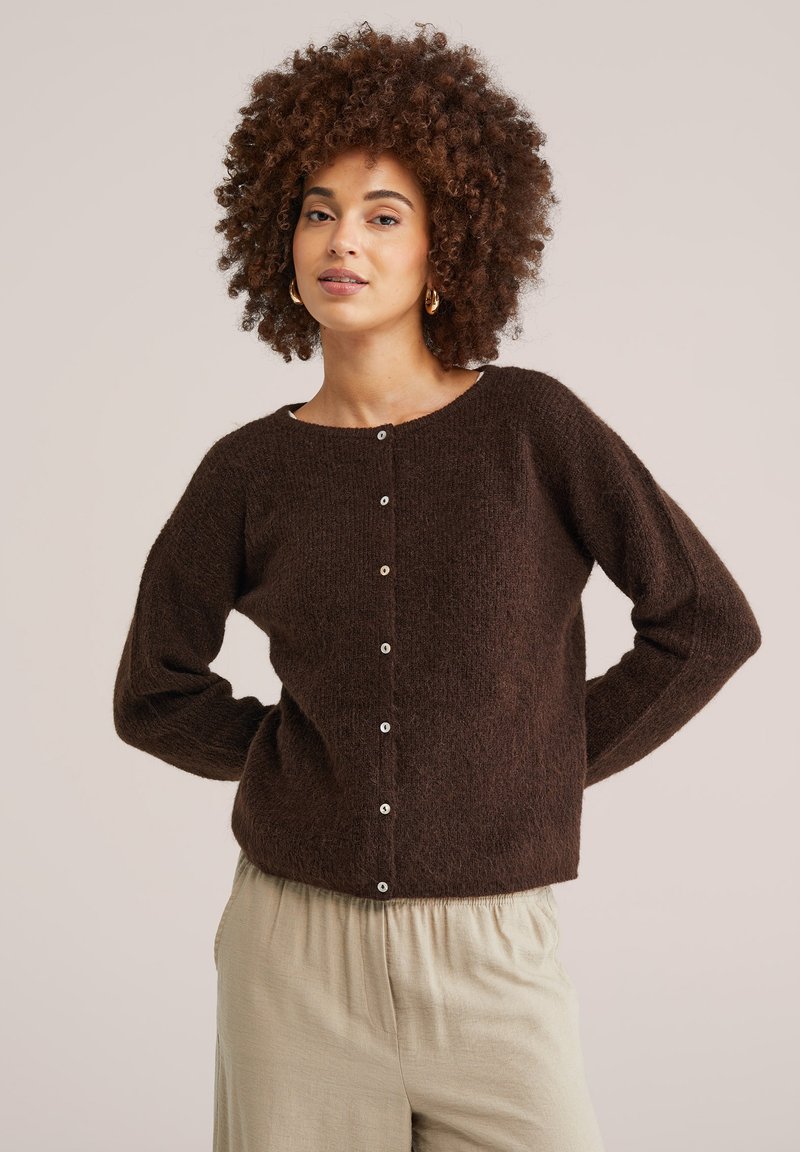 Sieviete ar cirtainiem matiem, valkā zelta stīpveida auskarus, tumši brūnu piepogātu kardiganu un gaiši beigzilas bikses, pozējot ar rokām aiz muguras.