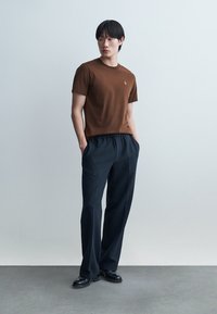 Mlad moški stoji z rokami v žepih, oblečen v rjavo srajco s kratkimi rokavi, črne široke hlače in črne čevlje, na enemobarvni svetli podlagi.