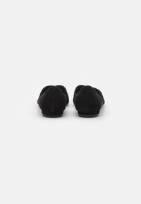 Sapatos slip-on pretos em camurça com um pequeno salto, biqueira arredondada e textura suave. O design apresenta uma silhueta minimalista e sem adornos visíveis.