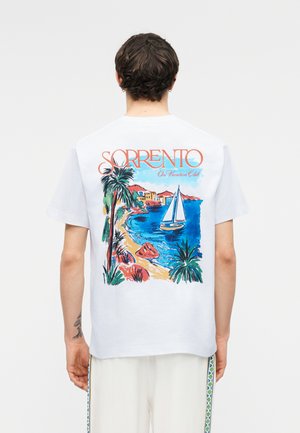 Moški, ki nosi belo majico s pisanim prizorom plaže Sorrento, palmami, jadralnim čolnom in napisom "SORRENTO On Vacation Club" na hrbtu.