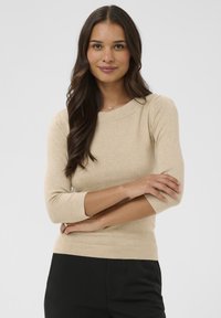 Haut beige ajusté à manches longues avec col rond et manches trois-quarts, confectionné en maille douce, porté avec un pantalon noir.